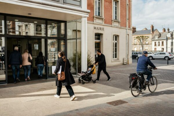 Des passants anonymes devant une mairie française, dans une scène de vie citoyenne en lumière naturelle.