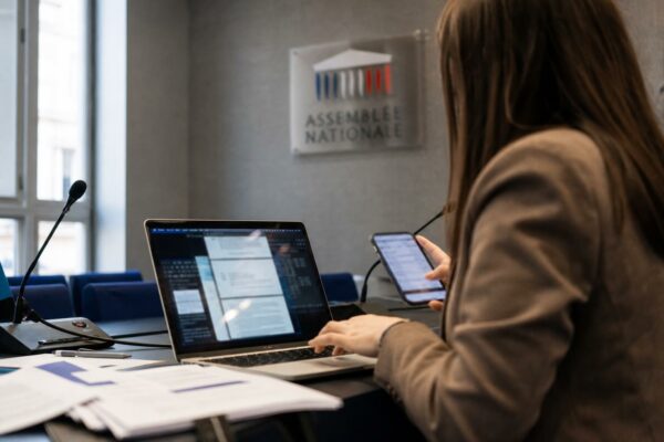 Journaliste dans une salle de presse parlementaire avec ordinateur et smartphone, ambiance de photojournalisme éditorial sur l’ingérence numérique.