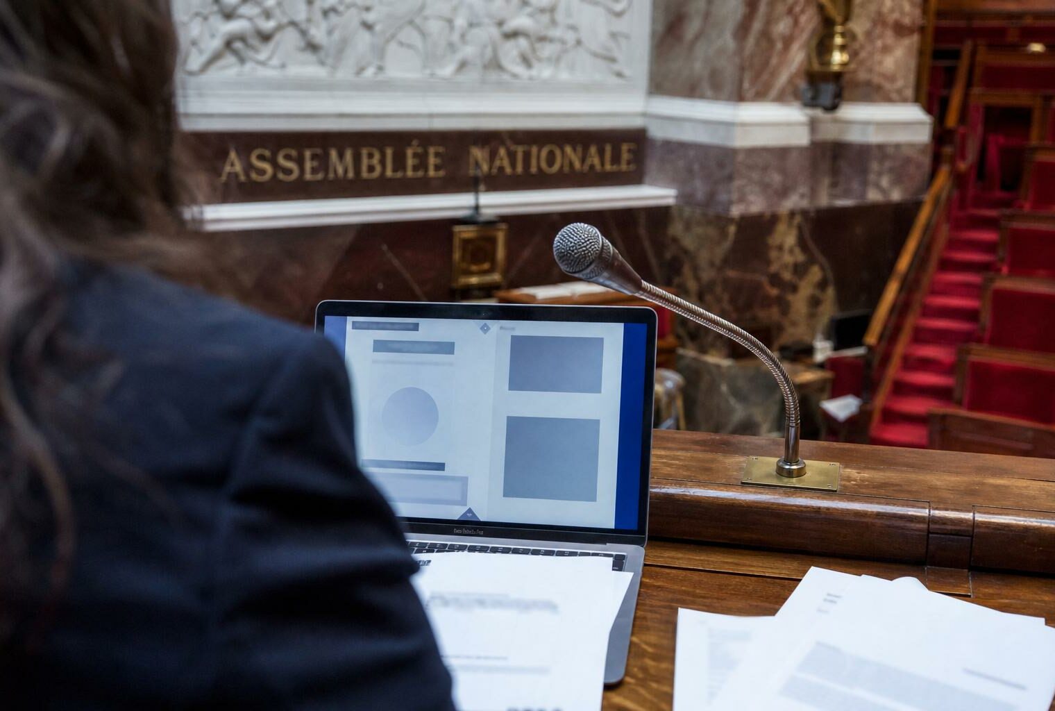 Vue documentaire d’un bureau parlementaire avec ordinateur, micro et hémicycle de l’Assemblée nationale en arrière-plan.