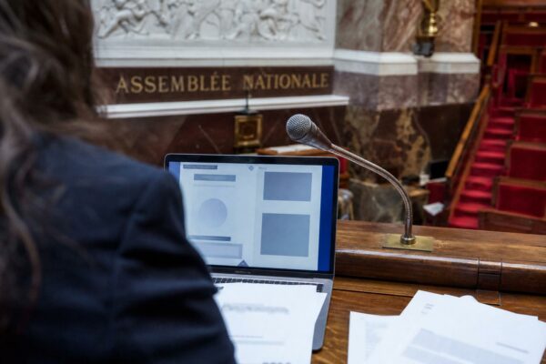 Vue documentaire d’un bureau parlementaire avec ordinateur, micro et hémicycle de l’Assemblée nationale en arrière-plan.