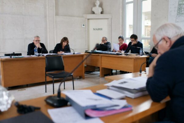 Salle municipale lumineuse avec micros, dossiers flous et chaise vide, scène de conseil local sur le logement.