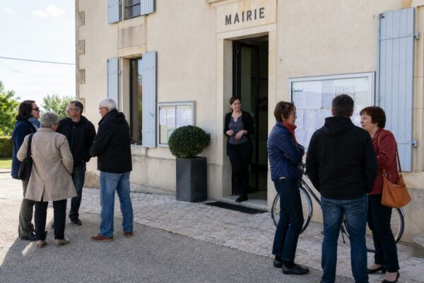 Des habitants anonymes sortent d’une mairie de village après une réunion locale, sous une lumière de jour claire.