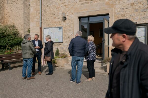 Des habitants et des élus locaux discutent devant une mairie de village en Aveyron, dans une lumière naturelle claire.