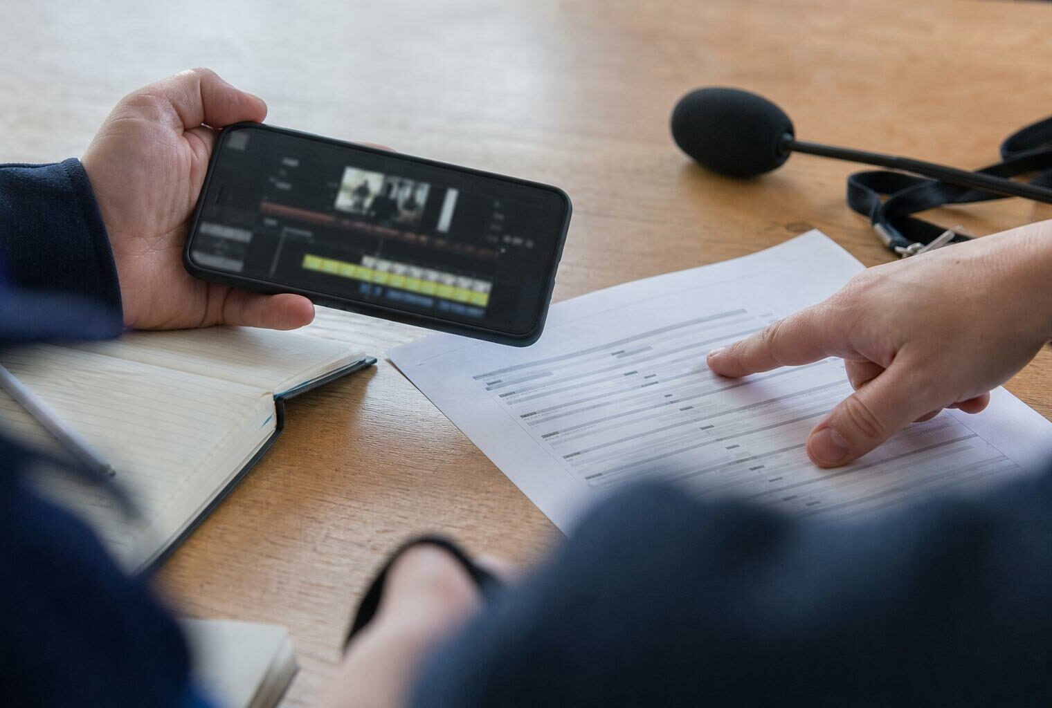 Main de journaliste et smartphone sur une table de rédaction, avec notes papier et micro sans logo.