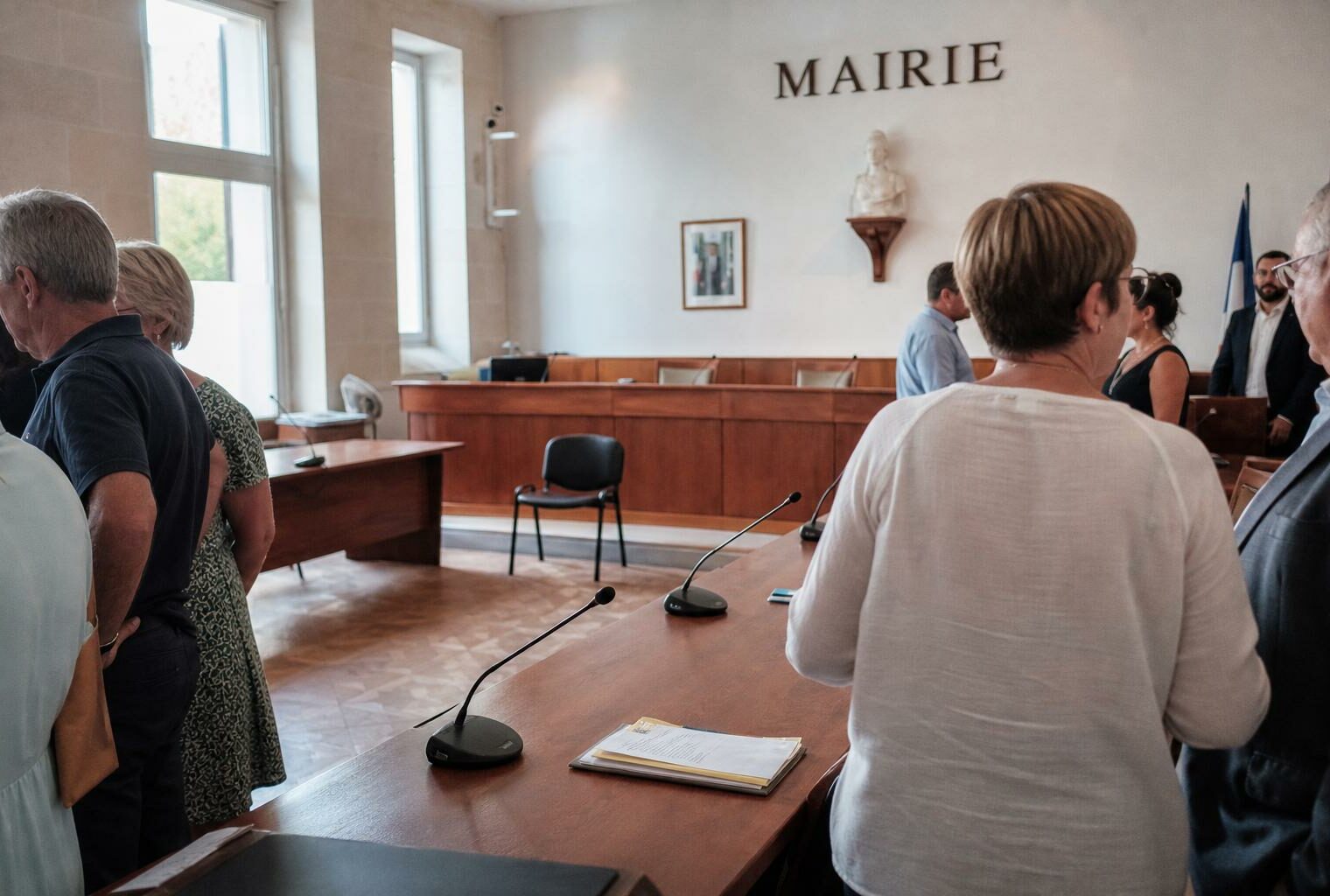Salle municipale française lumineuse avec micros, dossiers flous et chaise vide lors d’un rendez-vous politique.