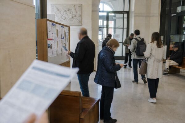 Des citoyens anonymes traversent un hall de mairie lumineux avec des affiches électorales floues en arrière-plan.