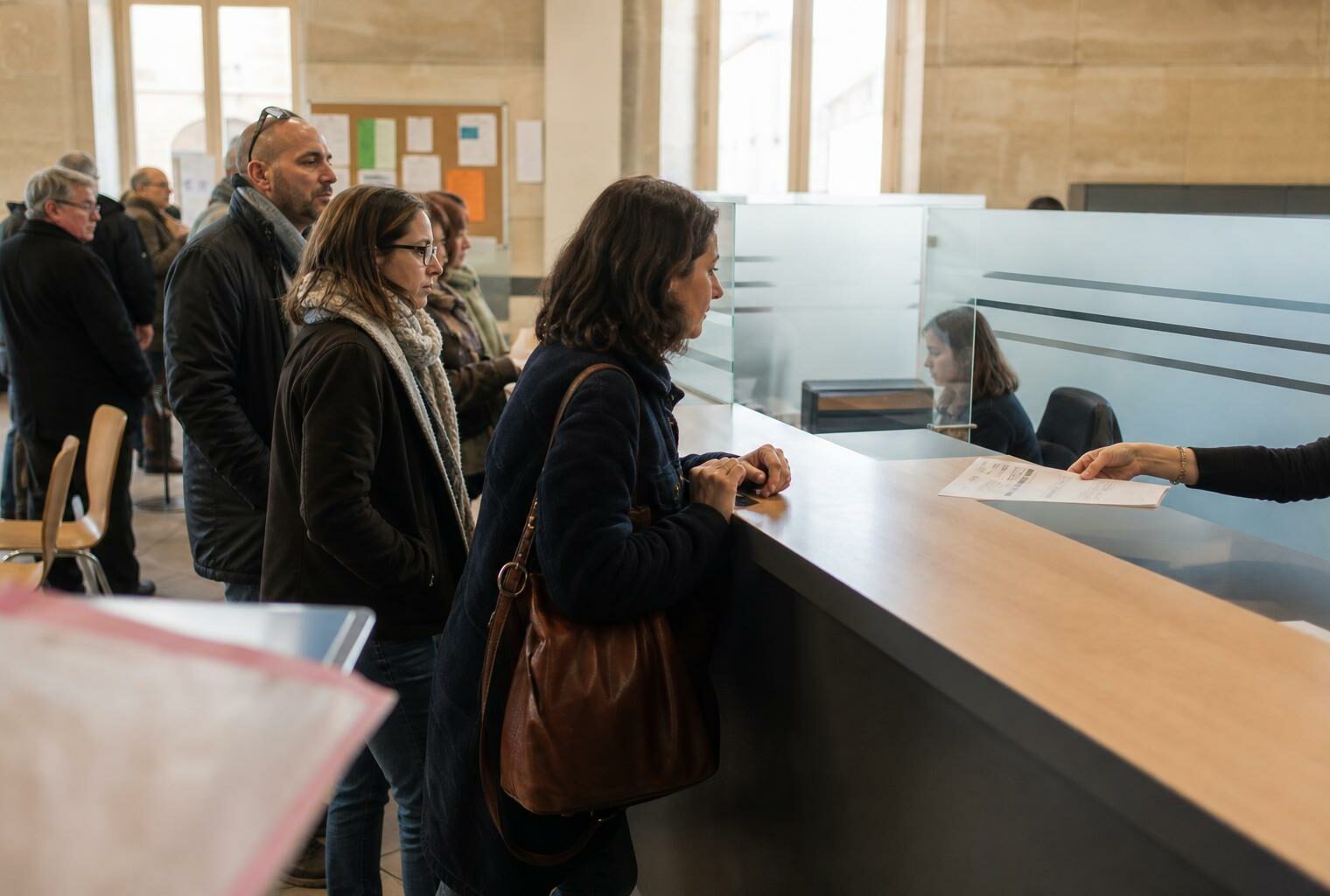 File d’attente dans un accueil de mairie, avec un dossier posé au guichet et des usagers anonymes.