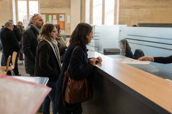 File d’attente dans un accueil de mairie, avec un dossier posé au guichet et des usagers anonymes.