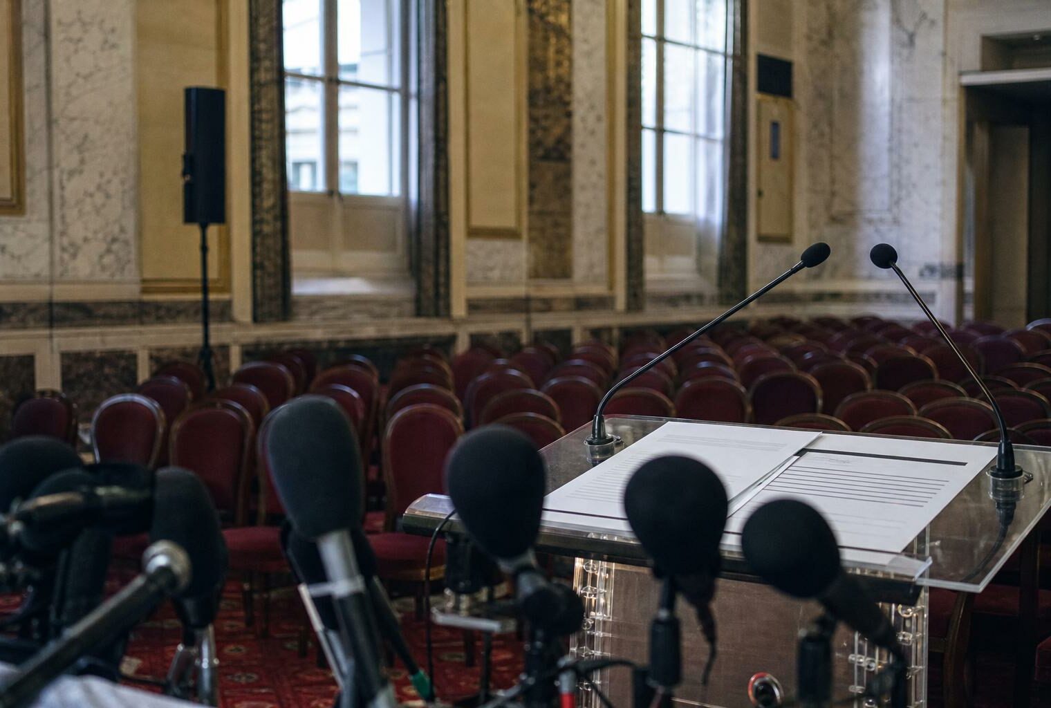 Salle de presse institutionnelle française vide avec pupitre, micros et rangées de sièges rouges, en style photojournalistique éditorial.