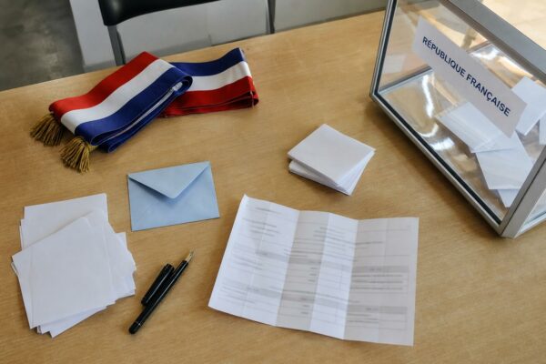 Flat lay photojournalistique d’un bureau de vote parisien avec bulletins, enveloppe bleue et urne transparente.