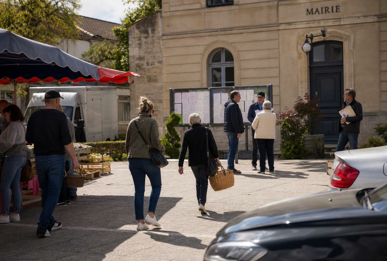 Des habitants anonymes traversent une place de ville moyenne près de la mairie et du marché, en lumière naturelle claire.