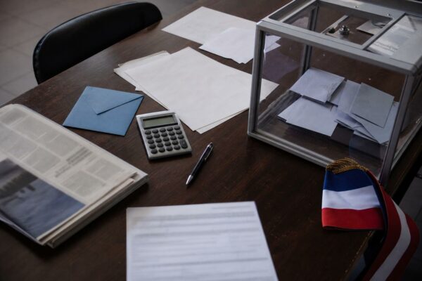 Vue en plongée d’une table de travail avec bulletins de vote, enveloppe, calculatrice et urne transparente, illustrant la présidentielle 2027 en contexte de crise.