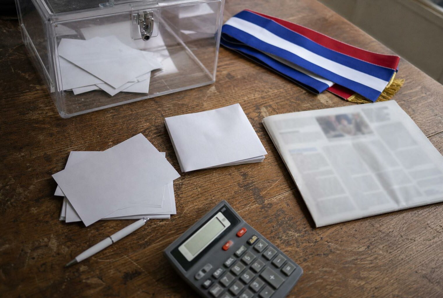 Vue plongeante d’un bureau de campagne politique français avec bulletins de vote, urne transparente et écharpe tricolore floue