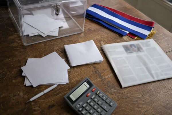 Vue plongeante d’un bureau de campagne politique français avec bulletins de vote, urne transparente et écharpe tricolore floue