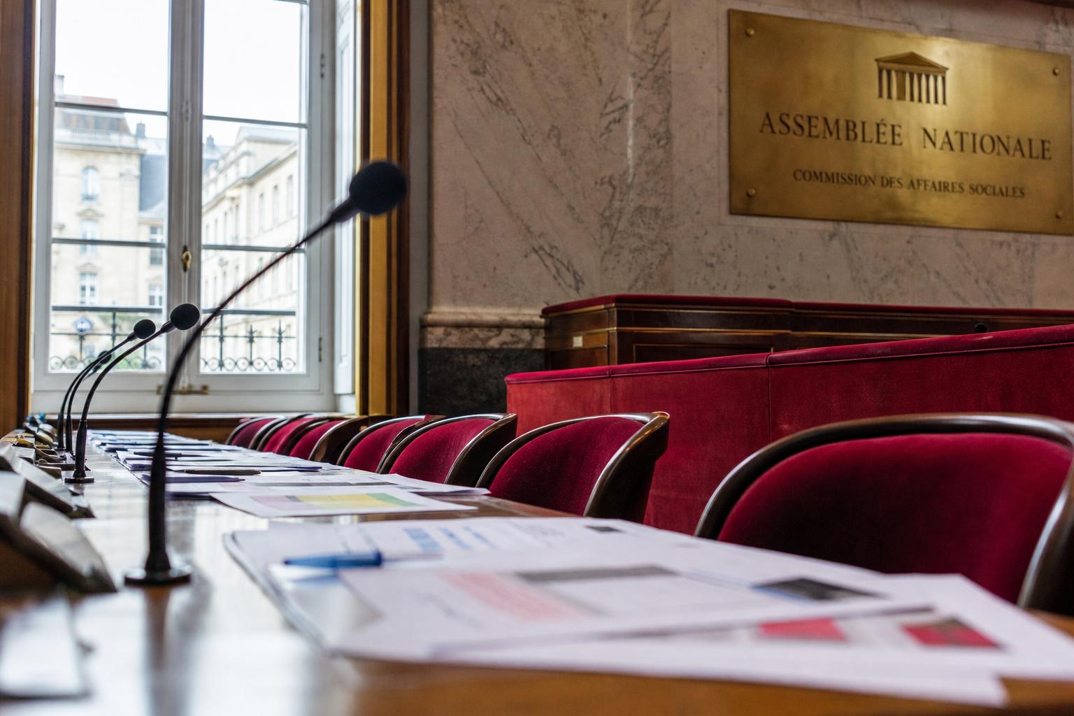 Salle de commission vide à l’Assemblée nationale avec micros et documents, illustrant le retard des décrets sur la prise en charge du cancer du sein