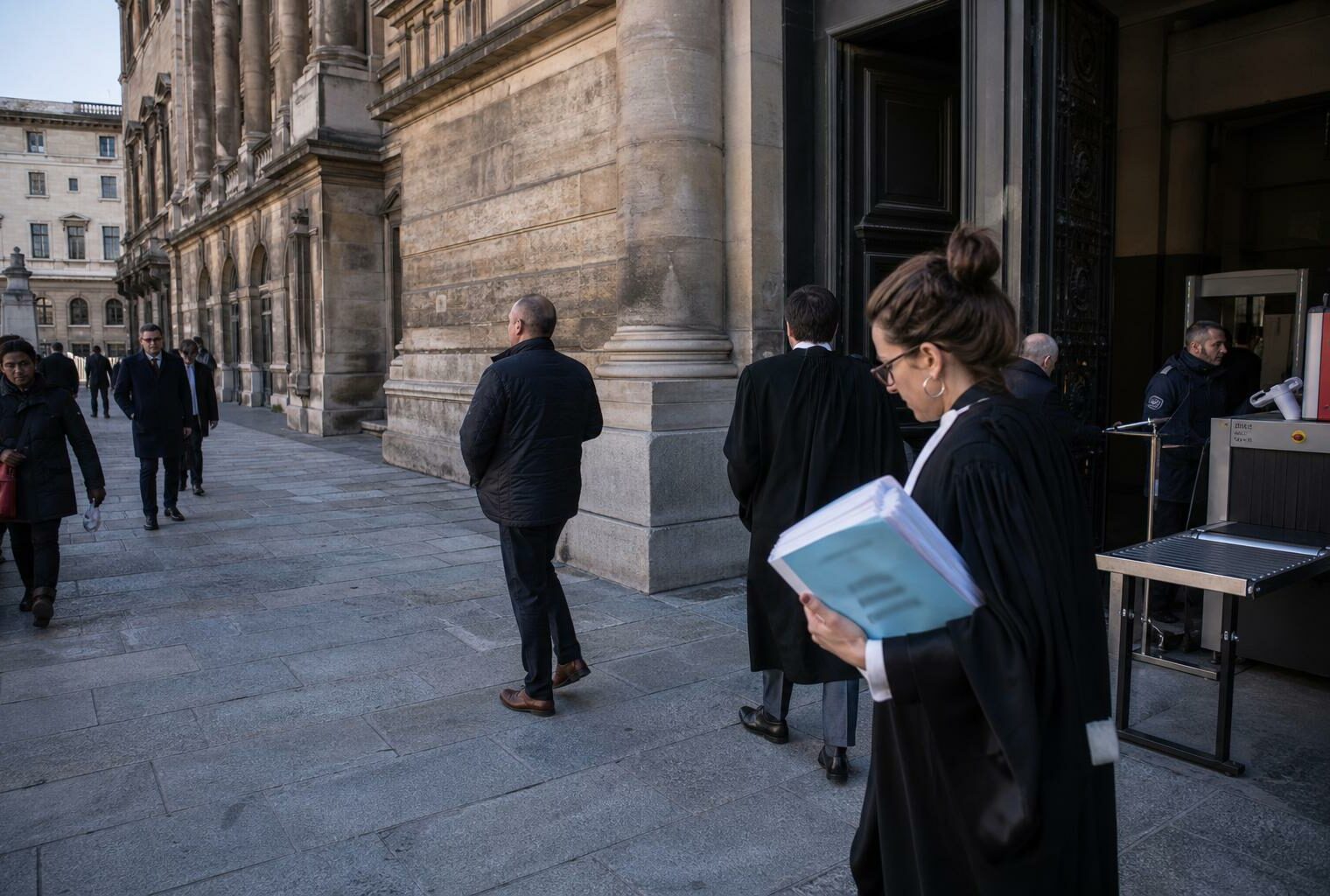 Devant un palais de justice parisien, des avocats et passants anonymes traversent une entrée de cour calme.
