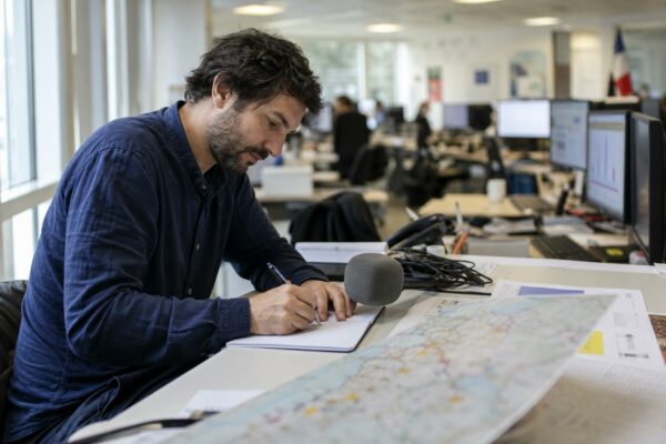 Journaliste en rédaction préparant un sujet économique, avec micro, carnet et écrans flous en lumière naturelle.