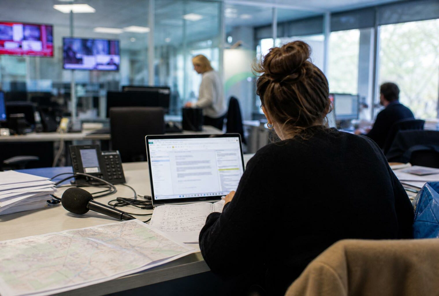Journaliste préparant un sujet territorial dans une rédaction française, avec carnet, micro sans logo et carte floue.
