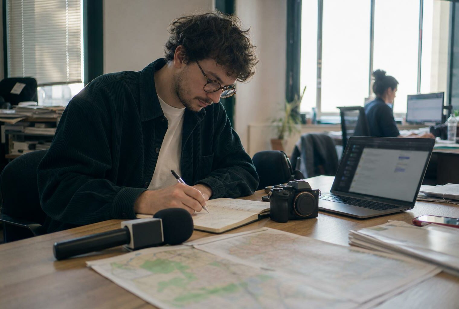 Journaliste préparant un sujet politique territorial dans une rédaction locale, avec carnet, micro et carte papier floue.