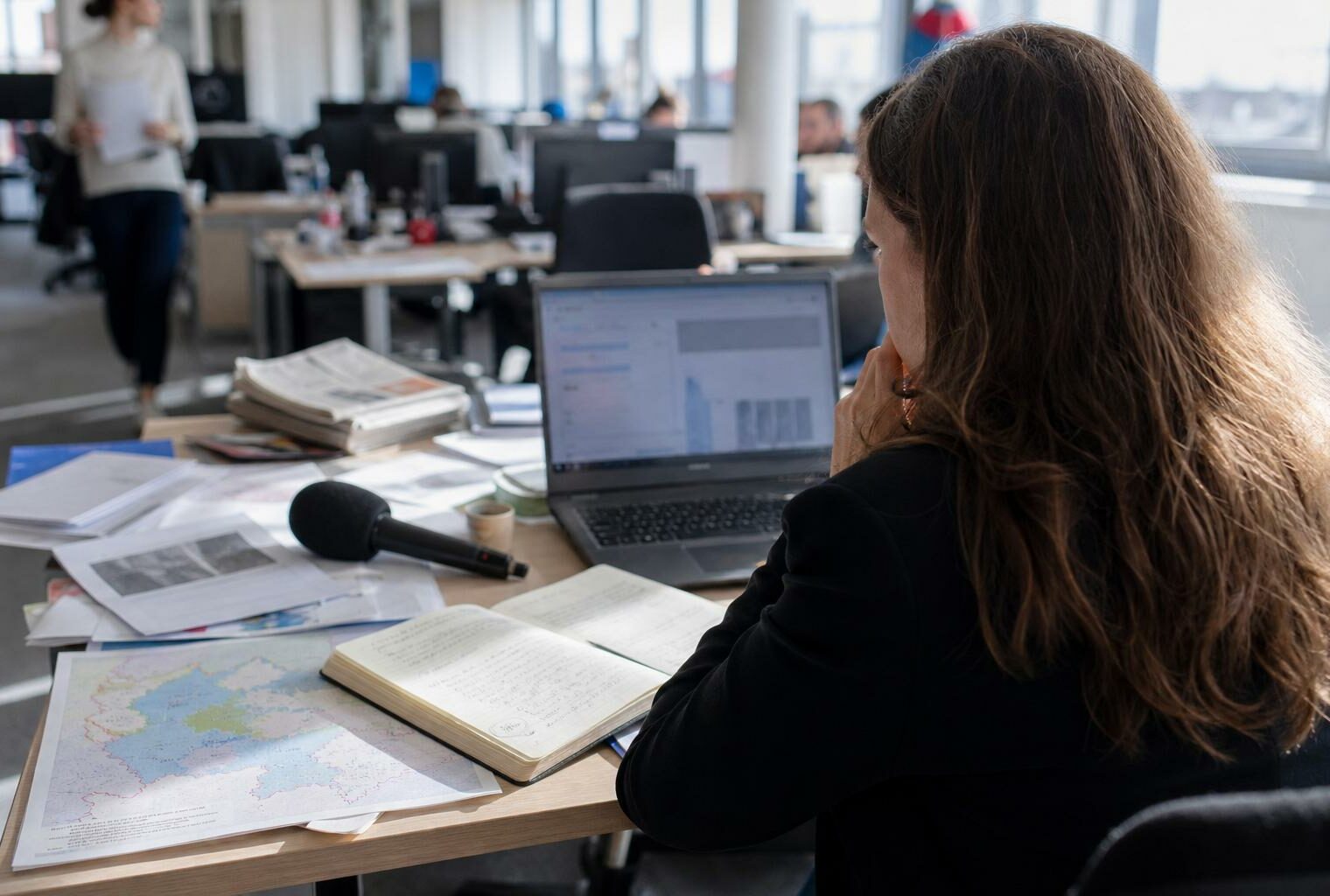 Journaliste en rédaction préparant un sujet territorial, avec carnet, micro sans logo et écran flou.