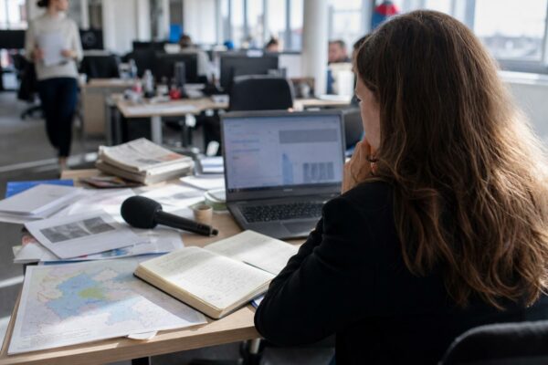 Journaliste en rédaction préparant un sujet territorial, avec carnet, micro sans logo et écran flou.