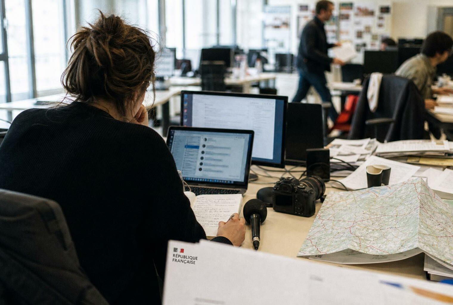 Journaliste en rédaction locale préparant un sujet politique avec carnet, micro sans logo et écrans flous.