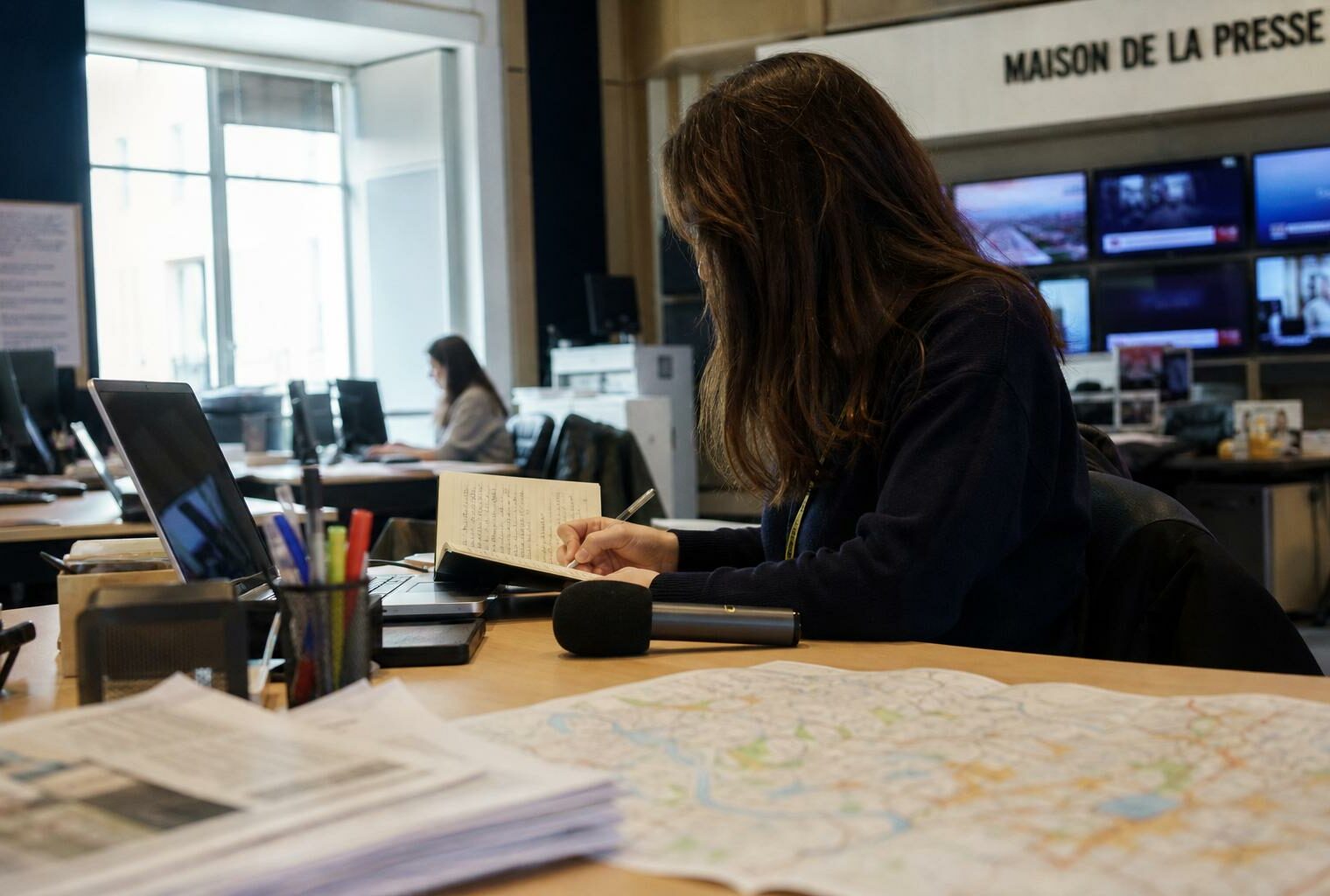 Un journaliste prépare un sujet territorial dans une rédaction française lumineuse, avec carnet, micro et écrans flous.