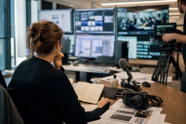 Journalistes dans une rédaction française, caméra et micros prêts pour un sujet politique, écrans flous en arrière-plan.