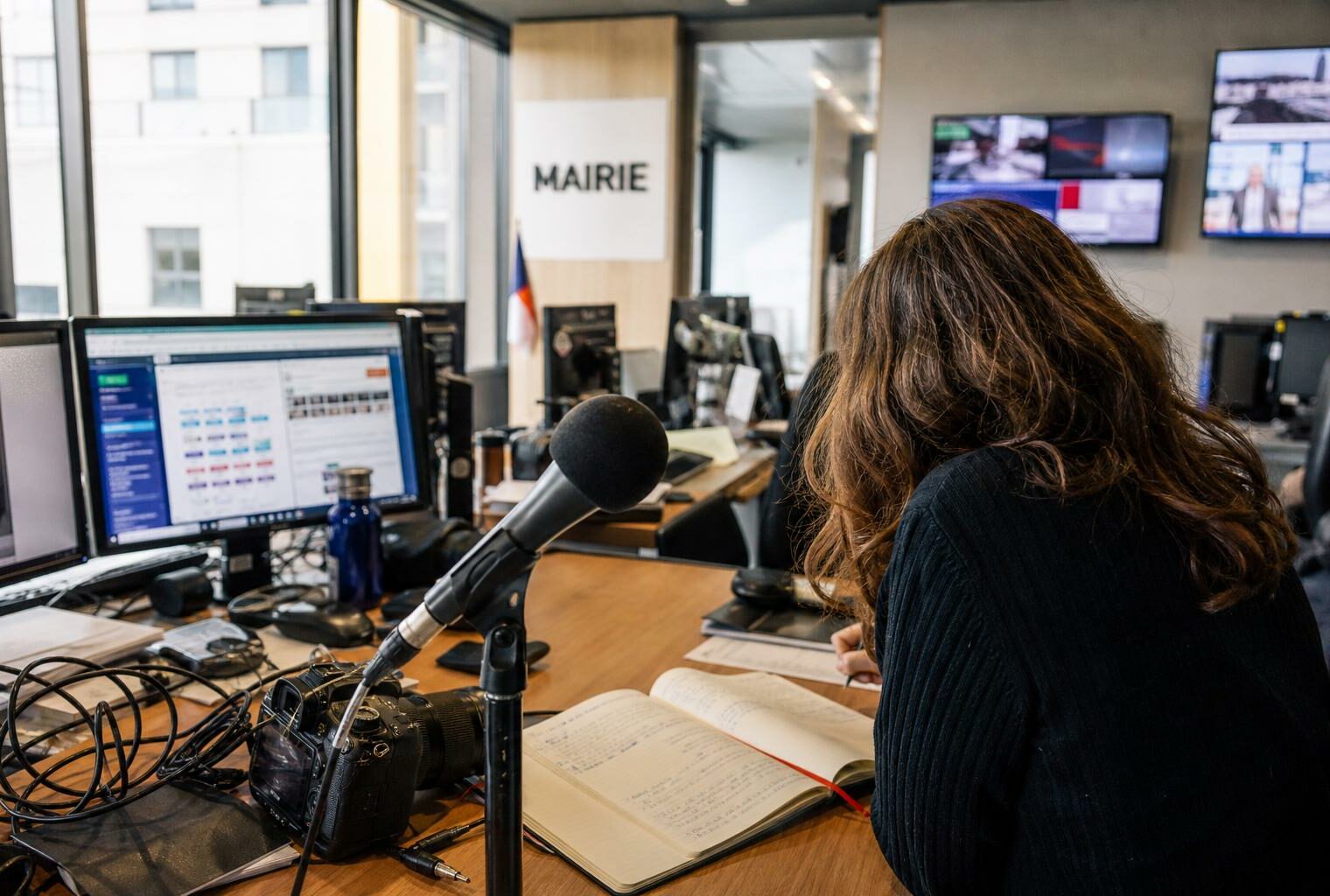 Une rédaction française prépare un sujet politique avec micro sans logo, carnet ouvert et écrans flous.