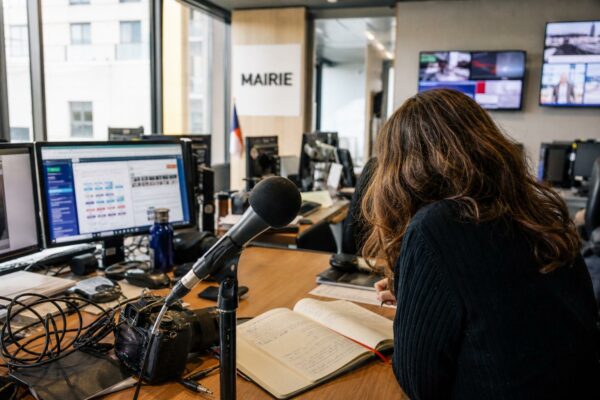 Une rédaction française prépare un sujet politique avec micro sans logo, carnet ouvert et écrans flous.