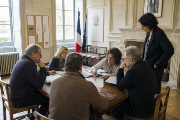 Réunion publique en mairie avec des habitants autour d’une table, sur fond de documents flous sur les retraites