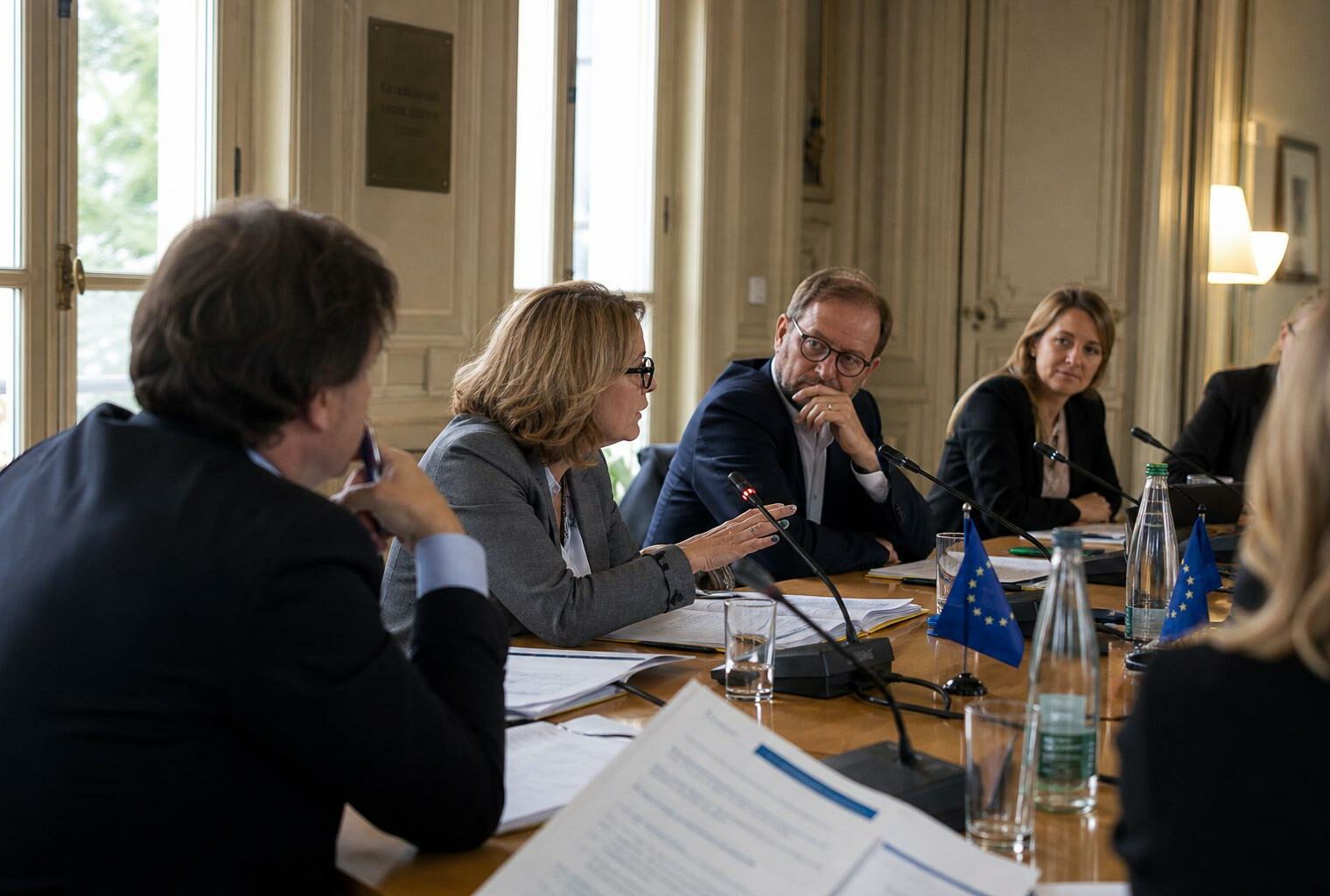 Réunion européenne sobre avec silhouettes de dos, dossiers, micros et petits drapeaux de table dans une salle claire.