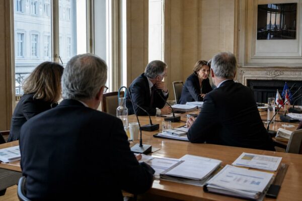 Réunion européenne sobre avec plusieurs silhouettes de dos, dossiers ouverts et drapeaux de table discrets.