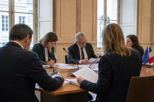 Réunion européenne sobre avec cinq silhouettes autour d’une table, dossiers et micros, lumière claire de salle institutionnelle.