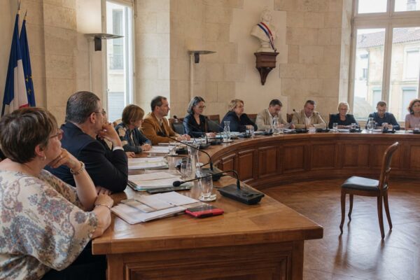 Salle municipale lumineuse avec chaise vide, micros et dossiers flous, ambiance de conseil local