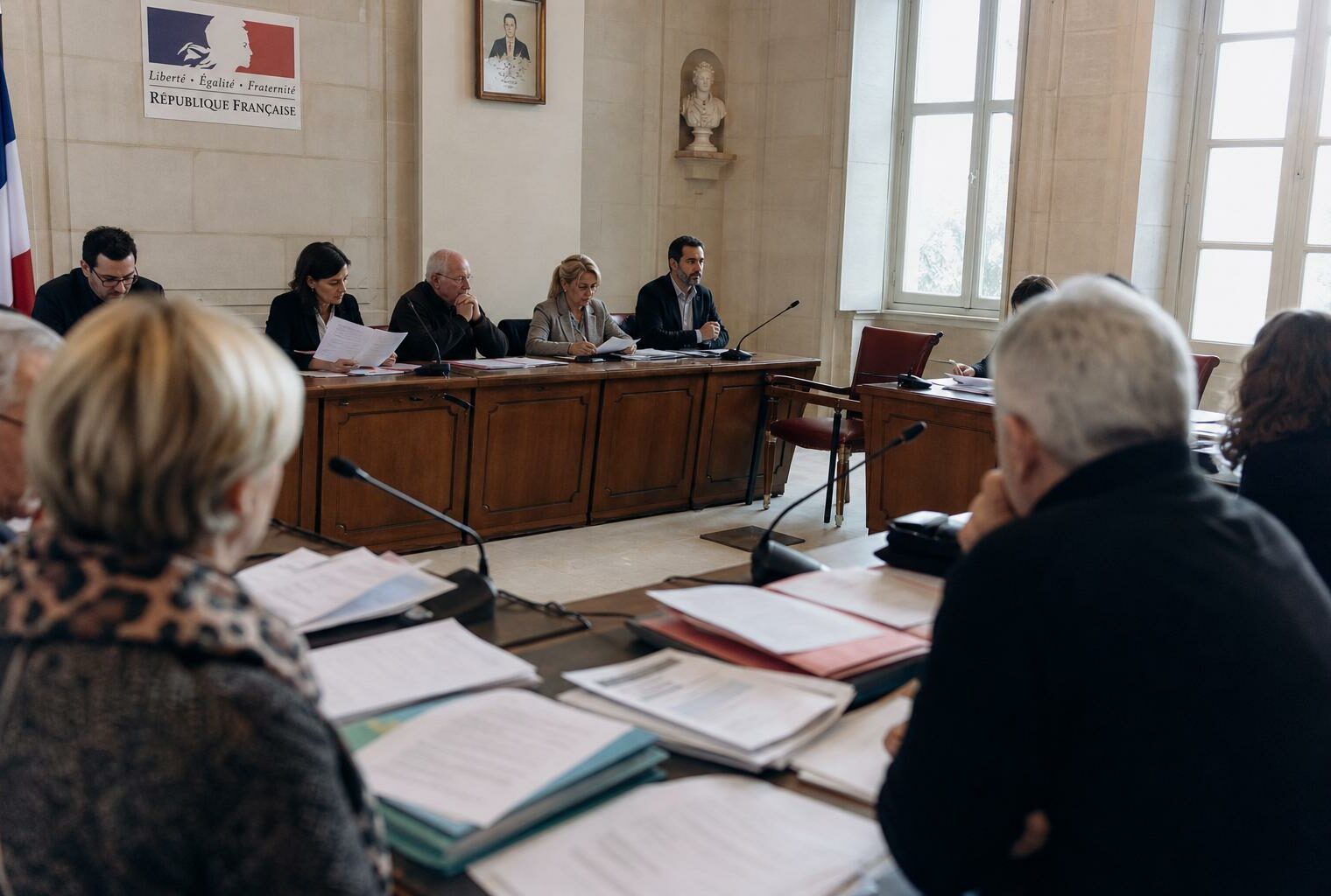 Salle municipale lumineuse avec chaise vide, micros de réunion et dossiers flous lors d’un conseil local