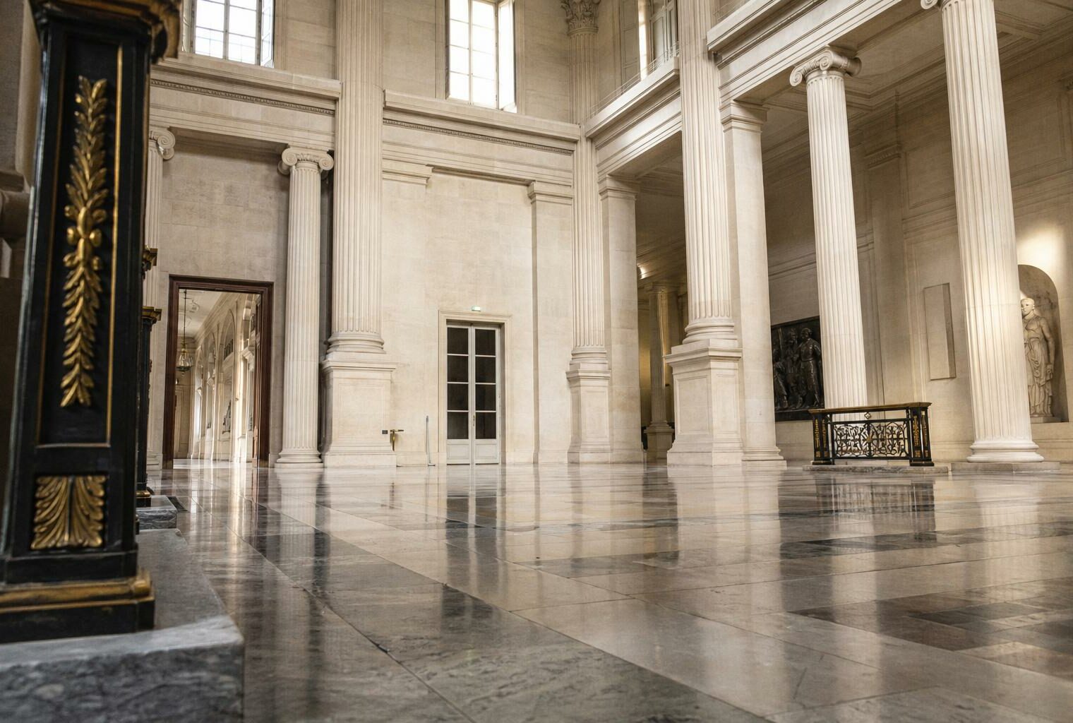 Salle vide de l’Assemblée nationale avec colonnes de pierre et lumière naturelle, vue architecturale en oblique.