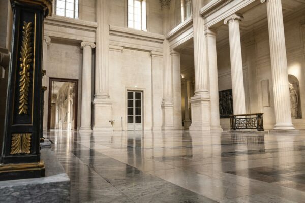 Salle vide de l’Assemblée nationale avec colonnes de pierre et lumière naturelle, vue architecturale en oblique.