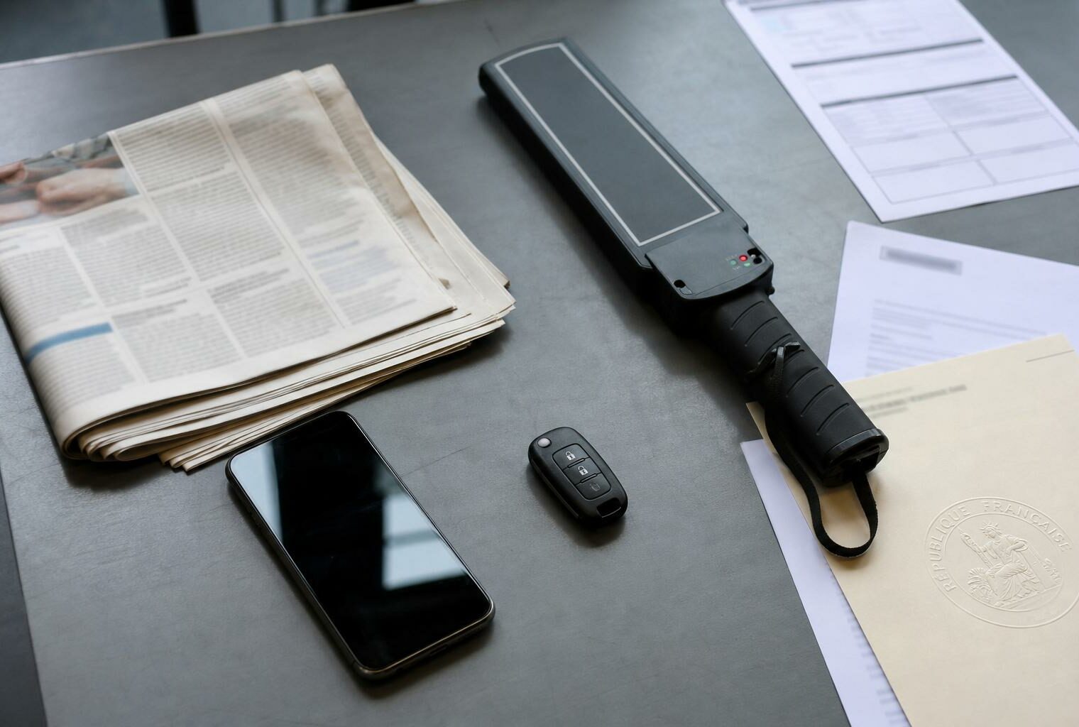 Vue de dessus d’un bureau avec journaux flous, smartphone éteint, détecteur d’examen et document officiel français, dans un style photojournalistique éditorial.