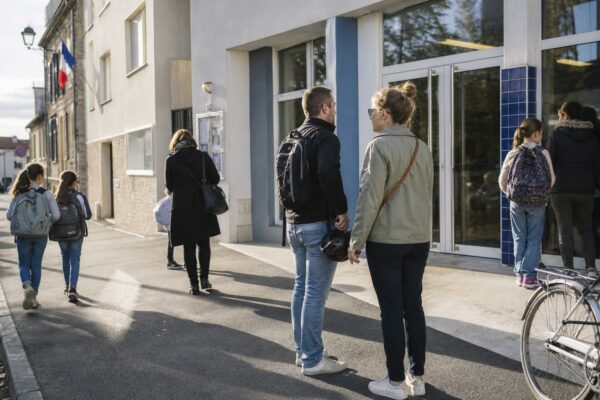 Parents anonymes à la sortie d’une école française, en matinée, devant une façade scolaire générique.