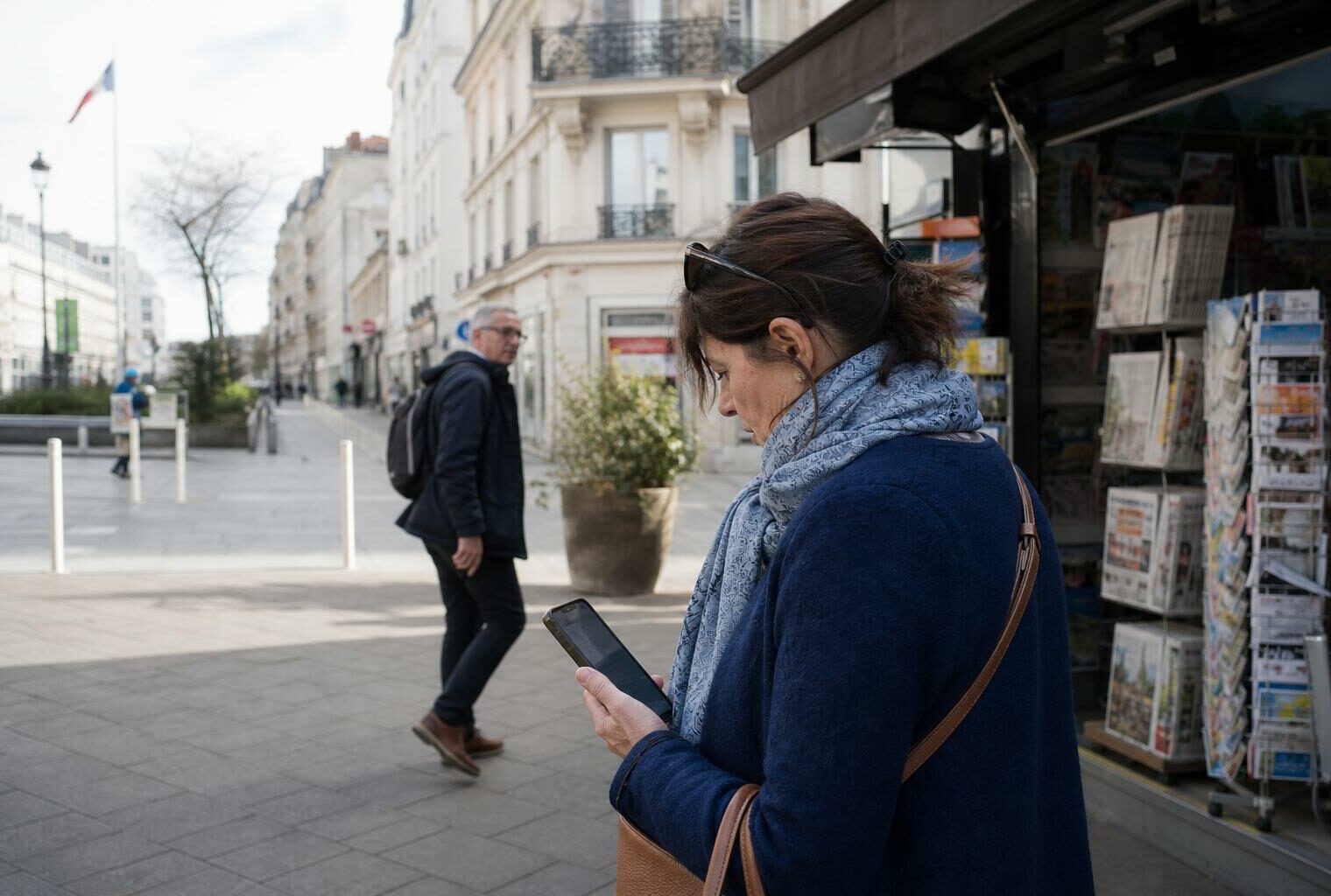 Un citoyen consulte son smartphone dans une rue française, près d’un commerce de quartier, en lumière du jour.