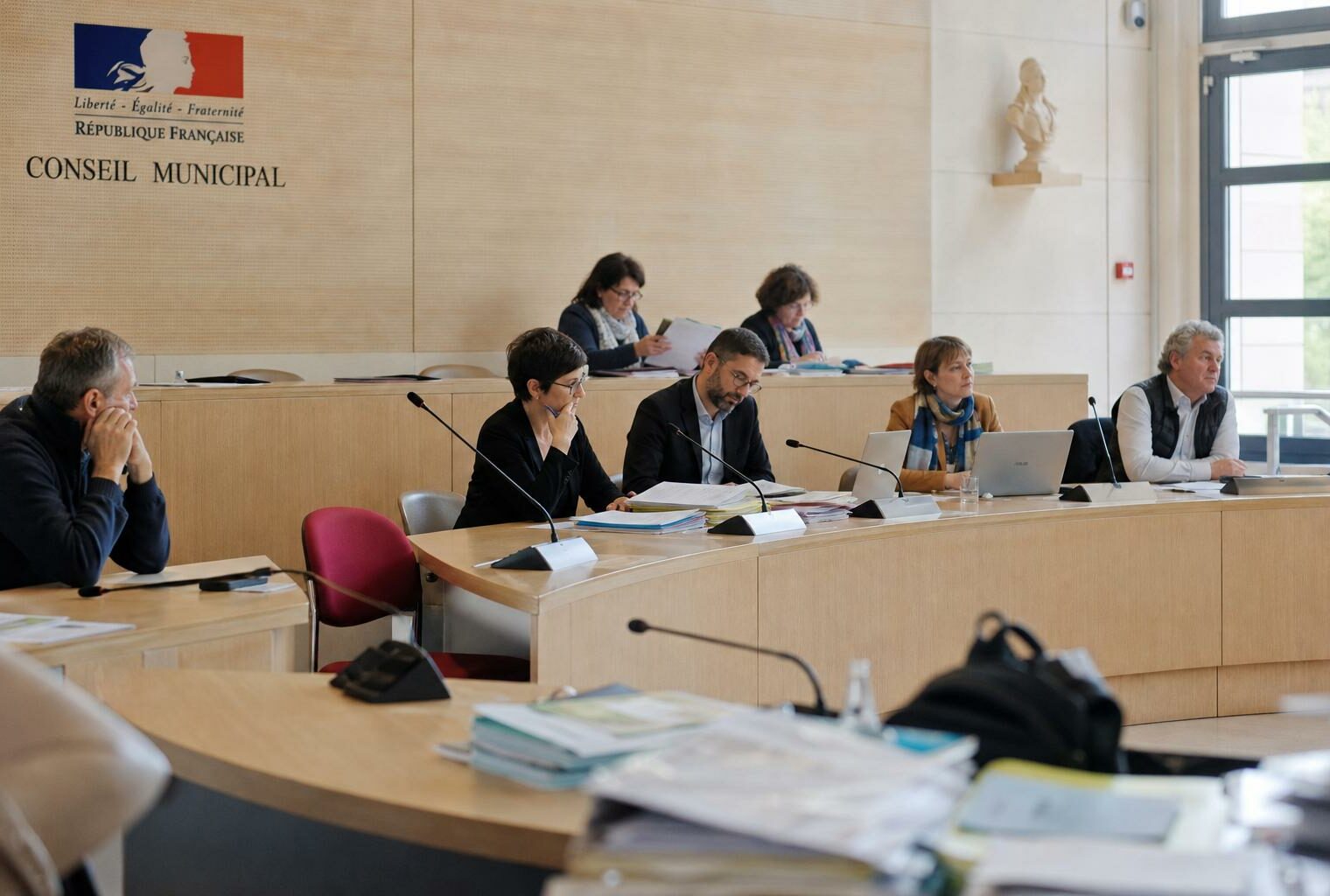 Salle municipale lumineuse avec chaise vide, micros et dossiers flous, ambiance politique sobre avant un départ des Bleus.