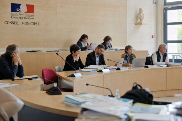 Salle municipale lumineuse avec chaise vide, micros et dossiers flous, ambiance politique sobre avant un départ des Bleus.