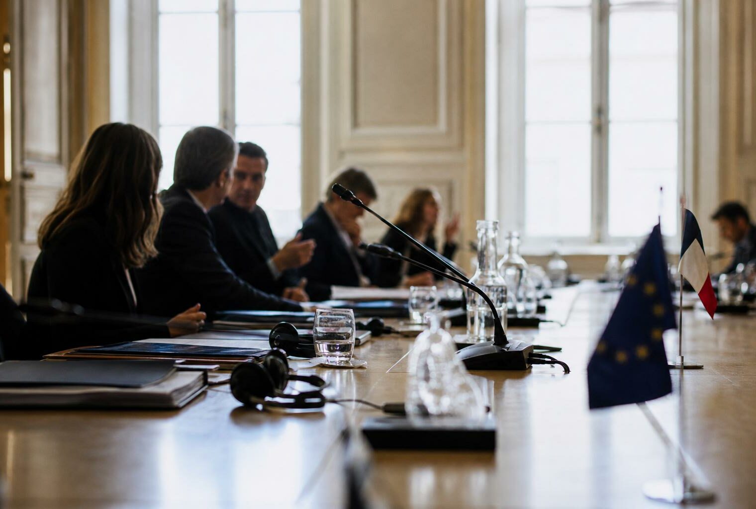Réunion institutionnelle avec dossiers, micros et silhouettes anonymes dans une salle claire de l’Assemblée nationale.