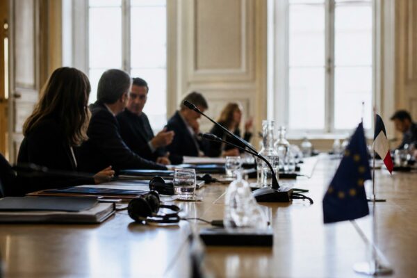 Réunion institutionnelle avec dossiers, micros et silhouettes anonymes dans une salle claire de l’Assemblée nationale.