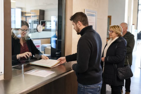 Accueil administratif lumineux dans une mairie, avec dossier posé au guichet et files de citoyens anonymes.