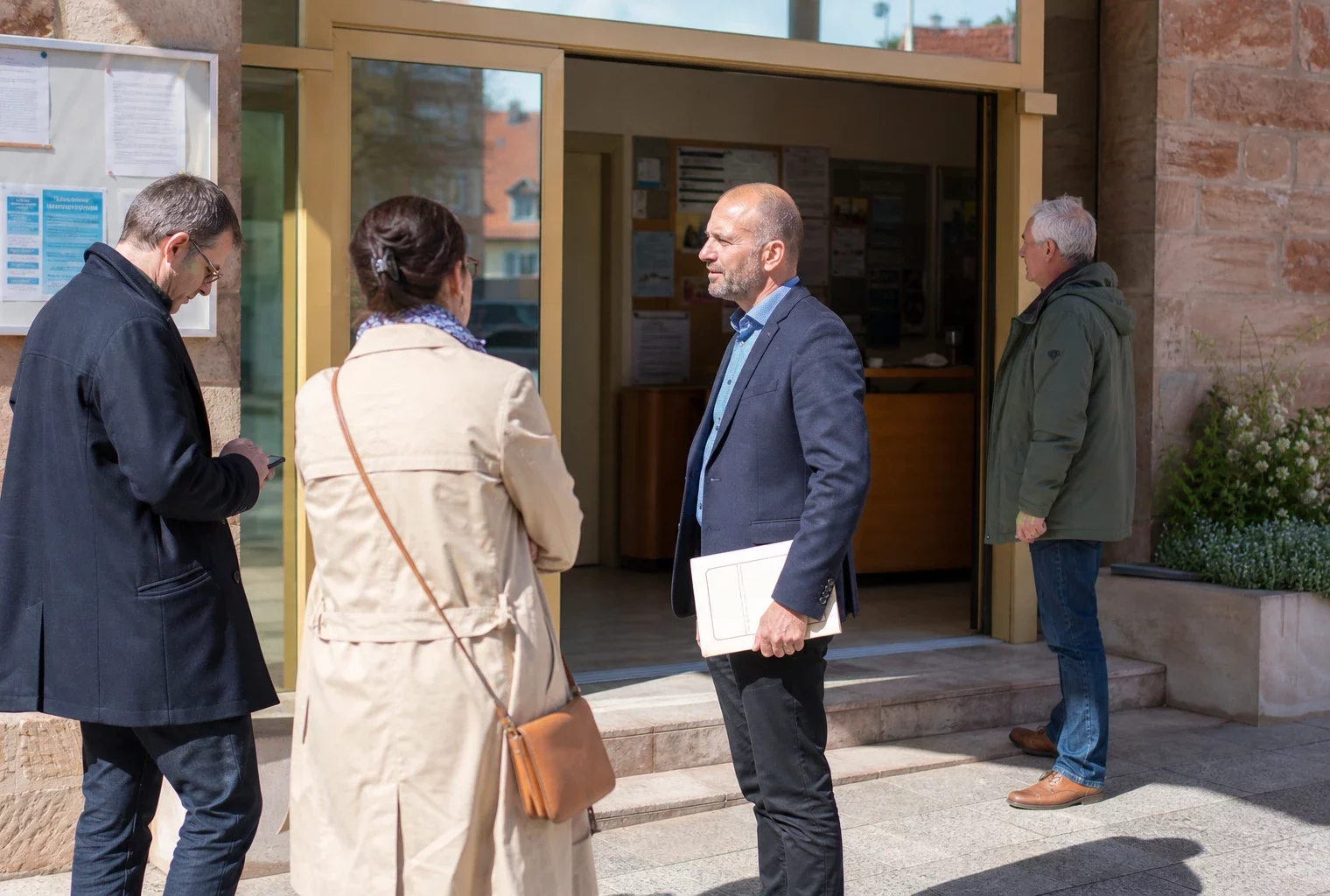 Entrée de mairie en Alsace avec citoyens anonymes et ambiance de service public local, en lumière naturelle.