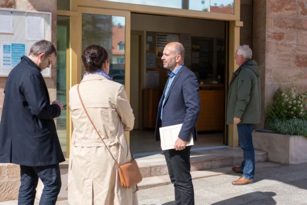 Entrée de mairie en Alsace avec citoyens anonymes et ambiance de service public local, en lumière naturelle.