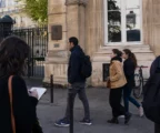 Scène de rue à Paris près d’un bâtiment diplomatique, avec passants anonymes et atmosphère journalistique calme.