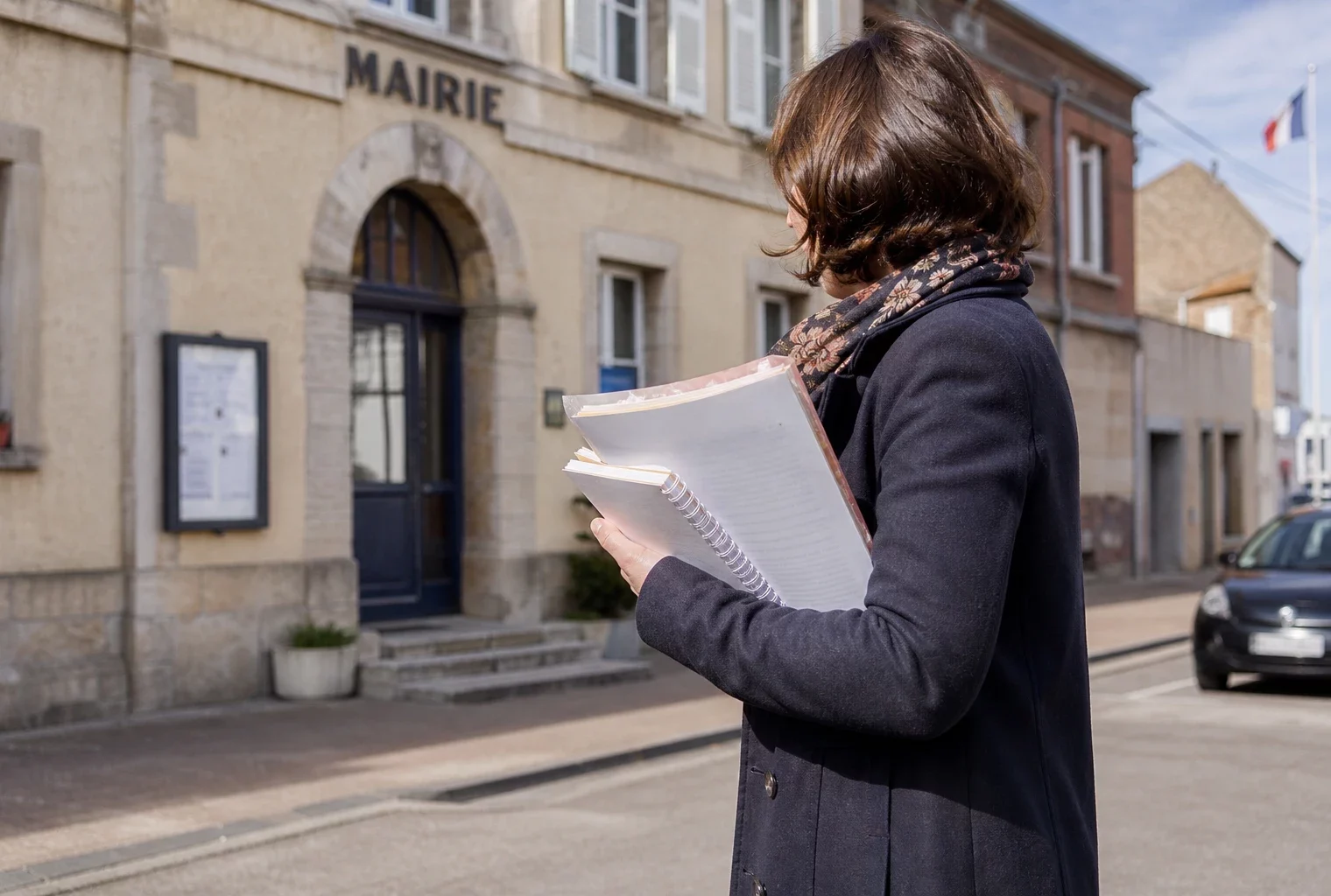 Un collaborateur municipal anonyme tient un dossier devant une mairie française, avec une rue calme en arrière-plan.